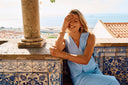 Frau in einem hellblauen Sommer-Outfit mit feinem Goldschmuck auf einer sonnigen Terrasse mit Meerblick. Die warmen Farben und die entspannte Atmosphäre sorgen für einen eleganten European-Summer-Look.