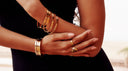 Nahaufnahme von goldenen Armreifen und Ringen an den Händen einer Frau in einem schwarzen Outfit. Eleganter Goldschmuck mit modernem und zeitlosem Design.