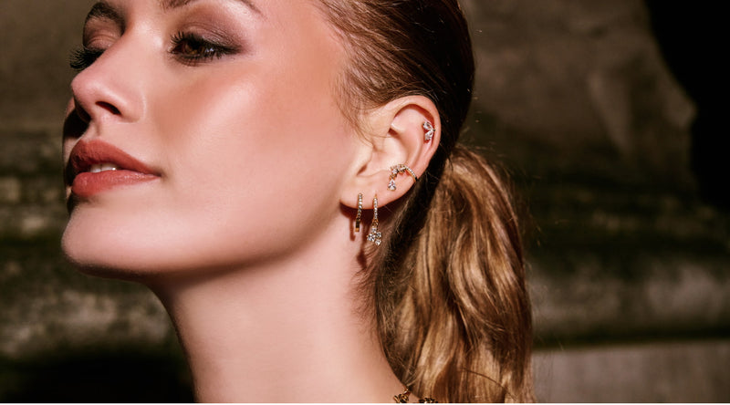 Nahaufnahme einer Frau mit mehreren goldenen Ohrringen und Piercings am Ohr. Eleganter Ohrschmuck mit funkelnden Steinen und modernem Ear Stack in warmem Licht.